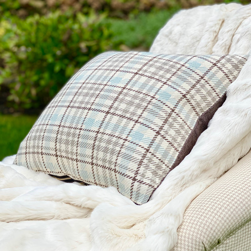The Coco Sky brown, cream and soft blue plaid square pillow rests on a creamywhite faux fur throw.