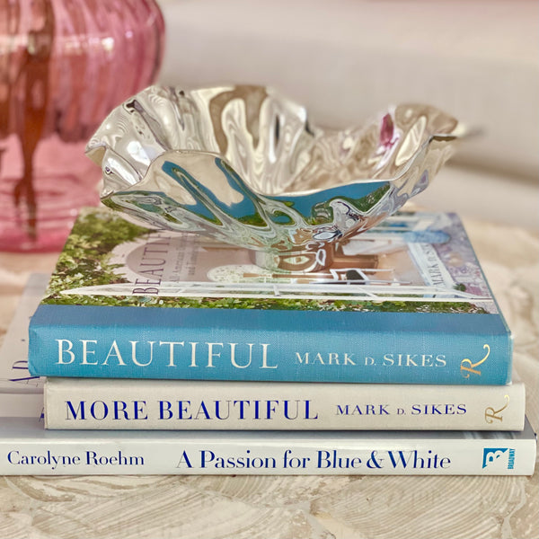 The Polished aluminum Vento Small Bloom Bowl by Beatriz Ball, features a wavy rippled edge. Shown resting atop a stack of design books.