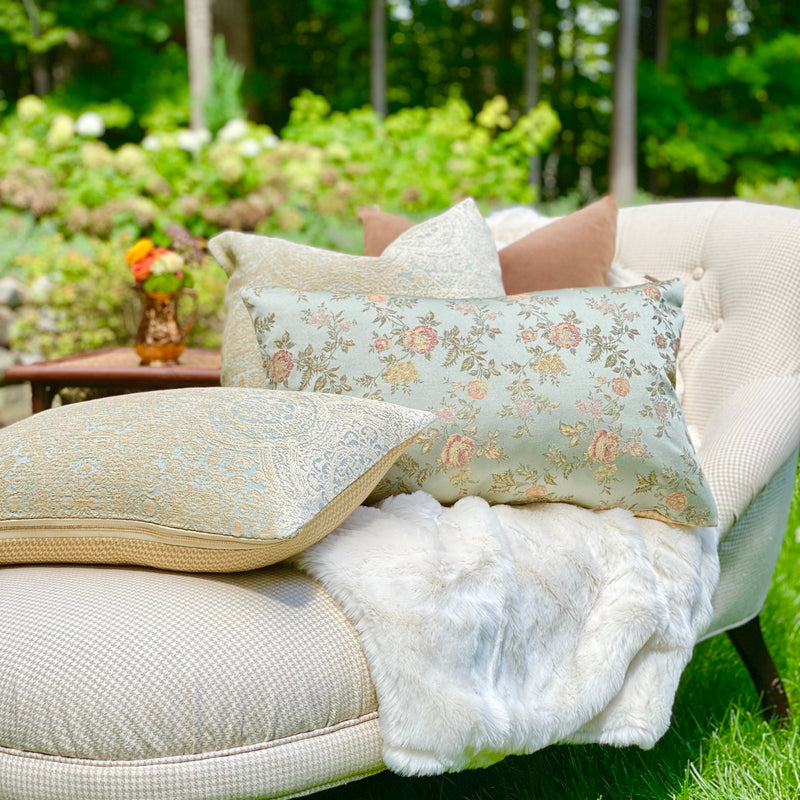 Large icy blue floral lumbar pillow with other pillows on a chaise outside with white faux fur throw.