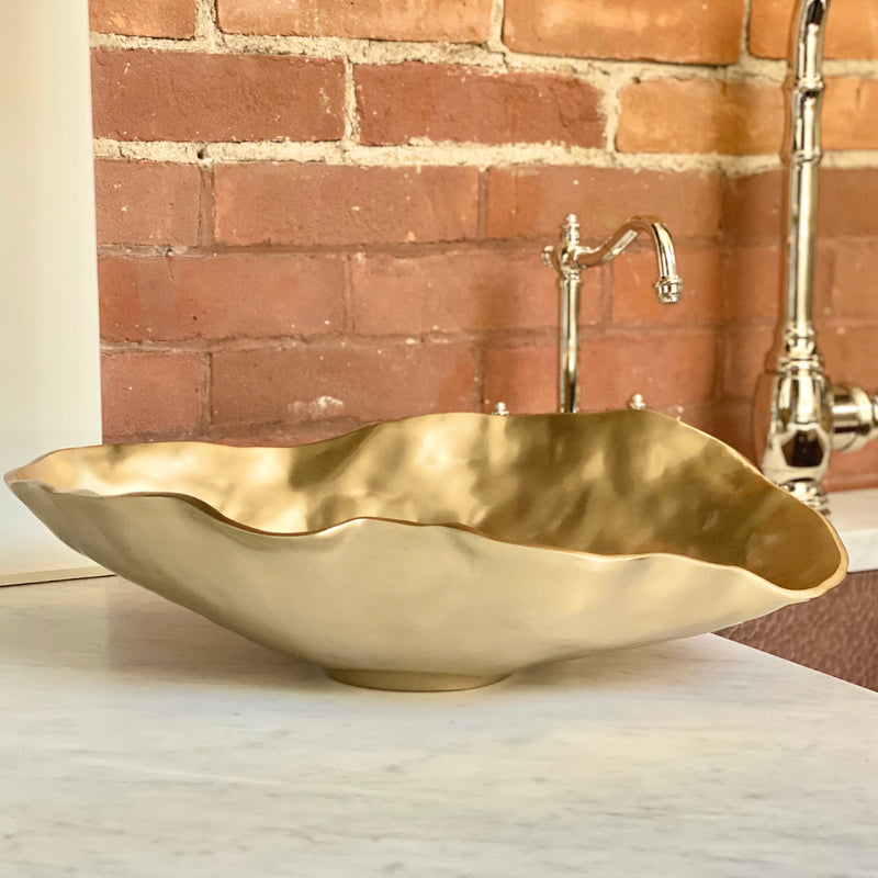 The large oval Maia Serving bowl with champagne gold finish, resting on a marrble counter with brick backsplash. From the Sierra Modern Collection, by Beatriz Ball.
